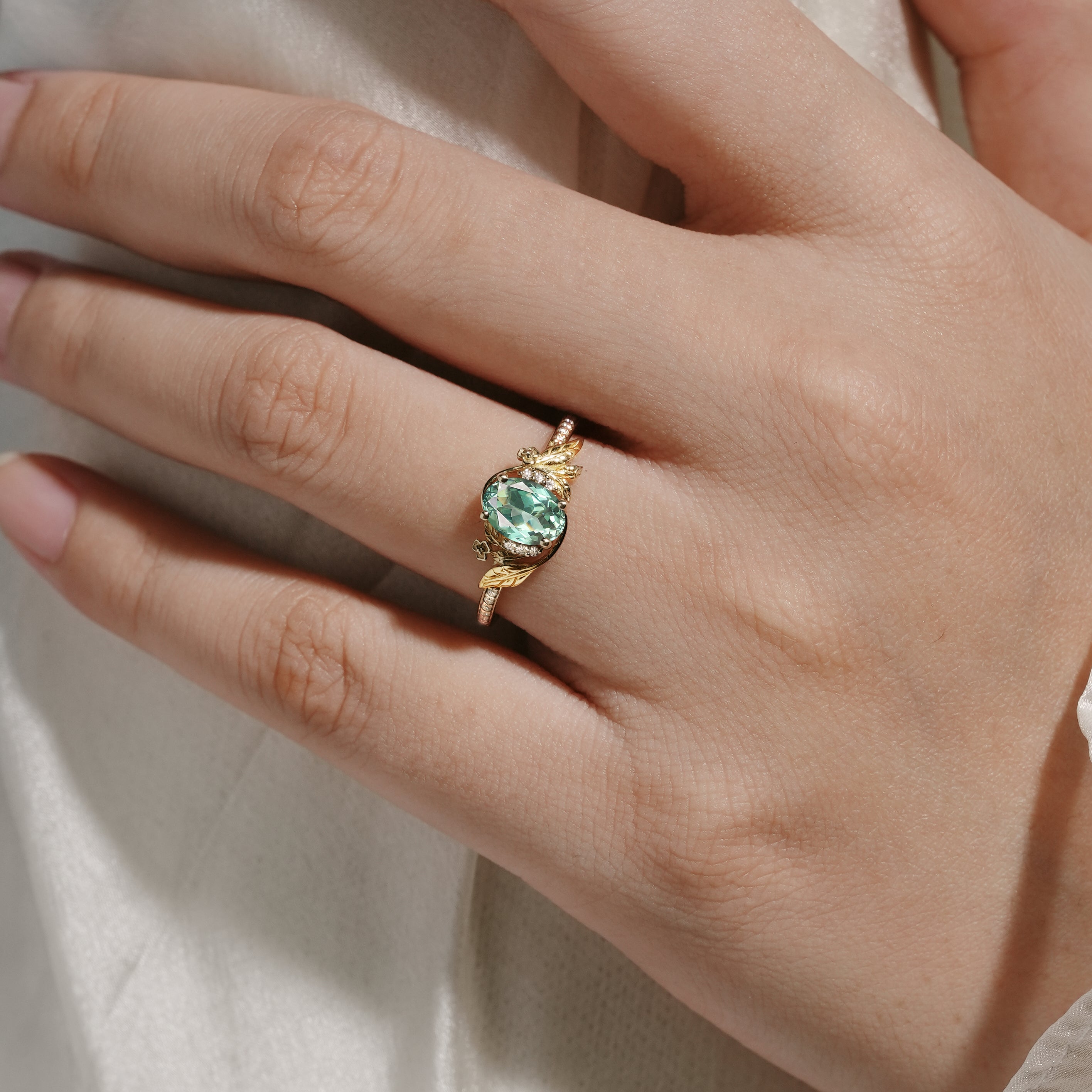 Gold engagement ring featuring an oval lab grown green sapphire with leaf and moissanite accents, worn on a hand.
