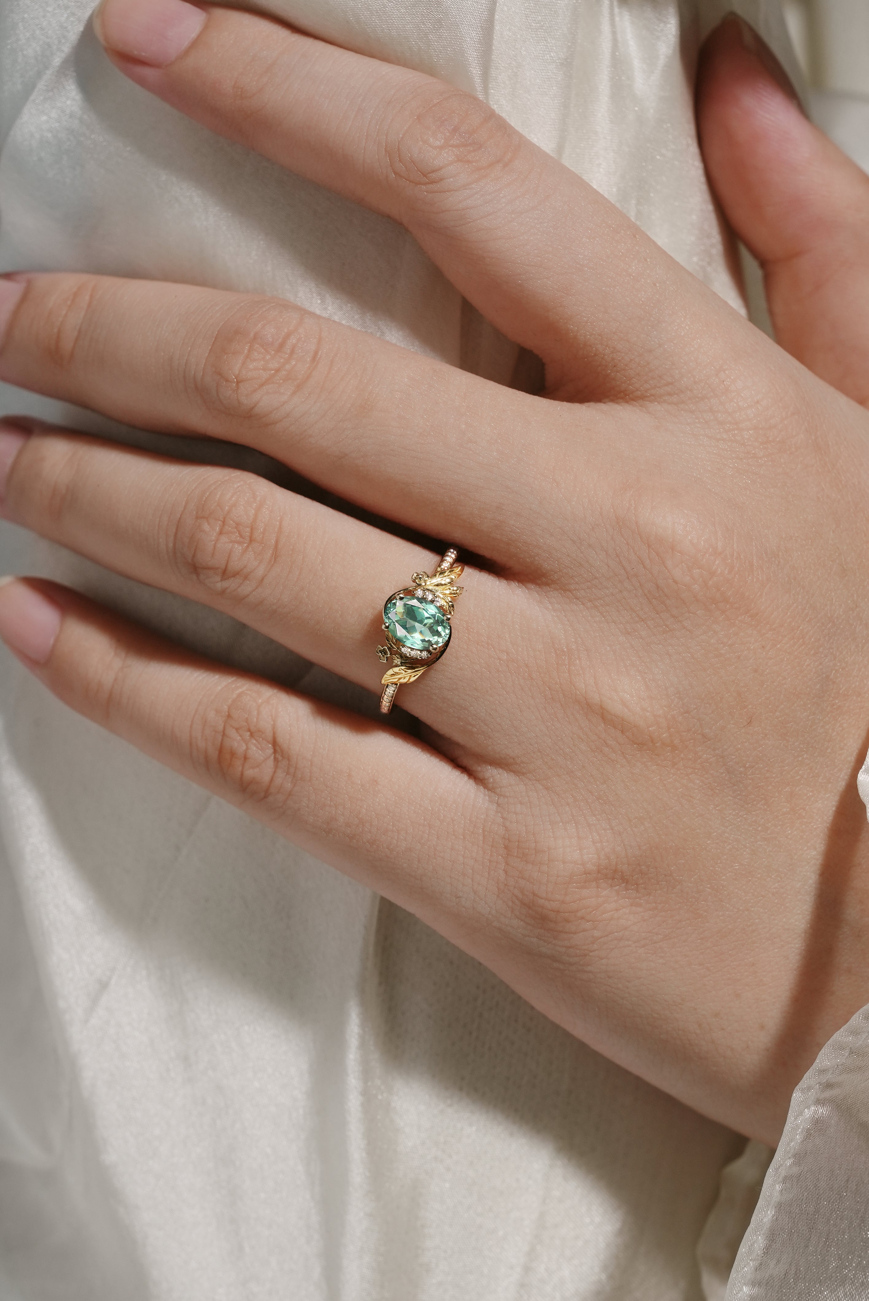 Gold engagement ring featuring an oval lab grown green sapphire with leaf and moissanite accents, worn on a hand.