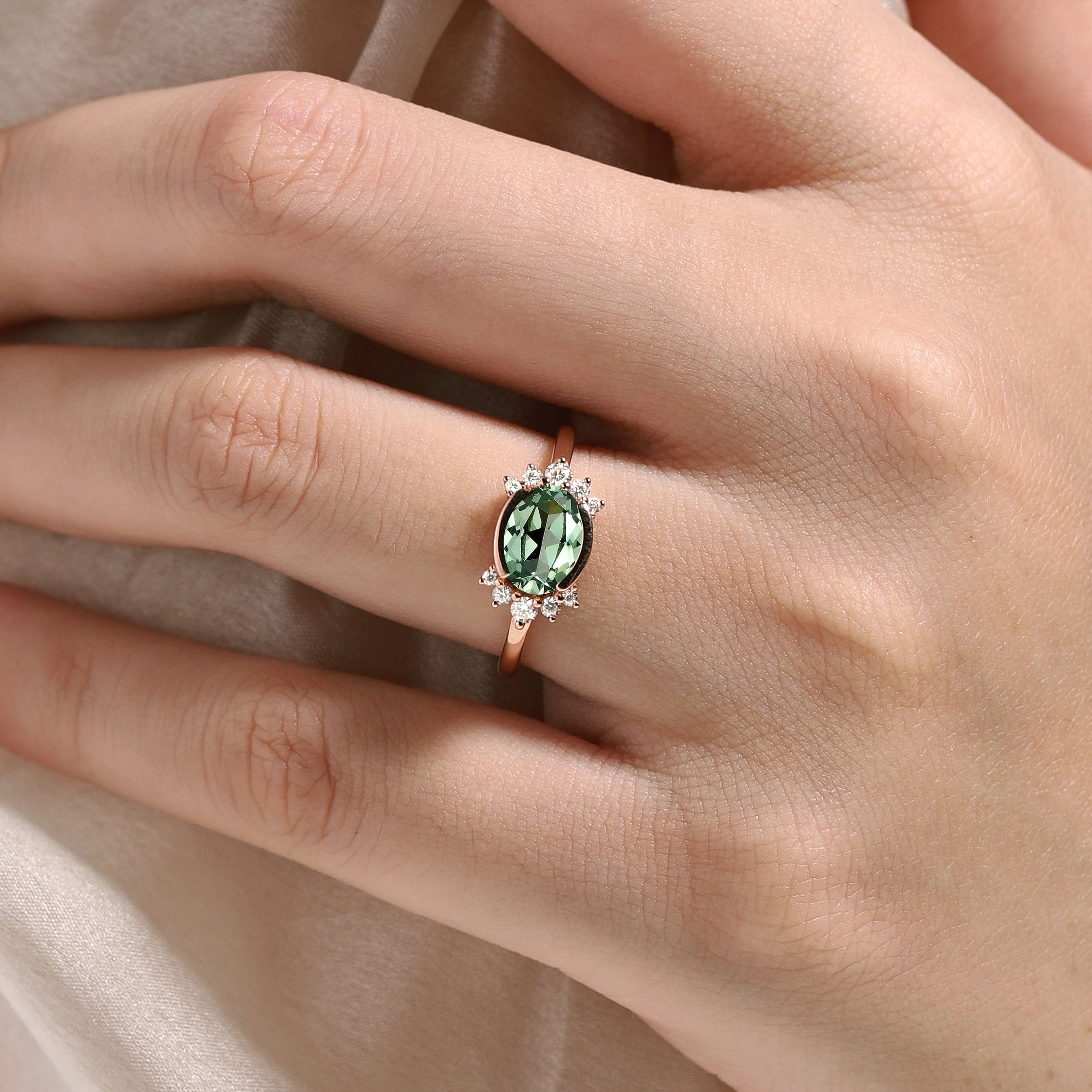 Rose gold oval green sapphire engagement ring with moissanite accents on a finger, bridal jewelry.