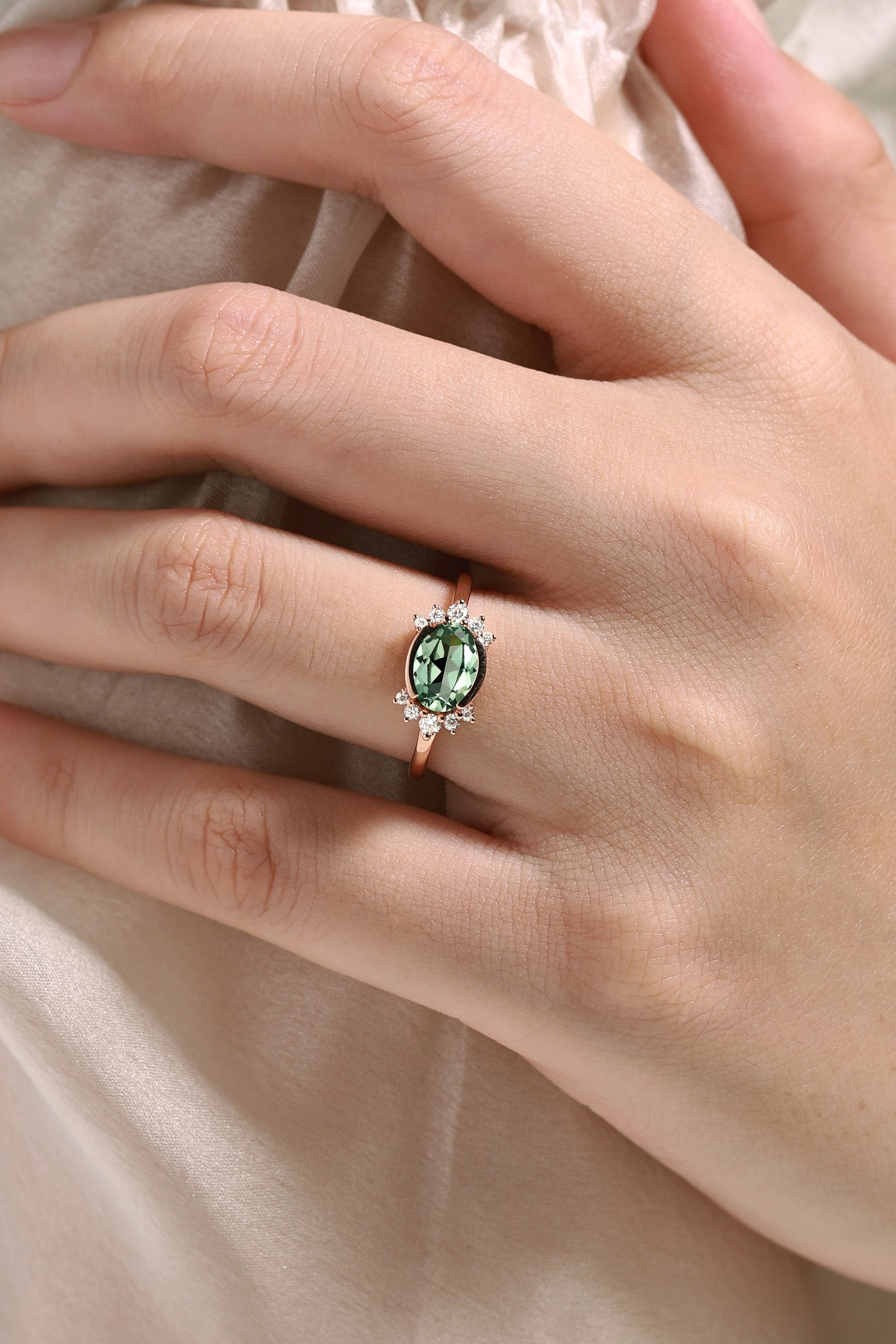 Rose gold oval green sapphire engagement ring with moissanite accents on a finger, bridal jewelry.