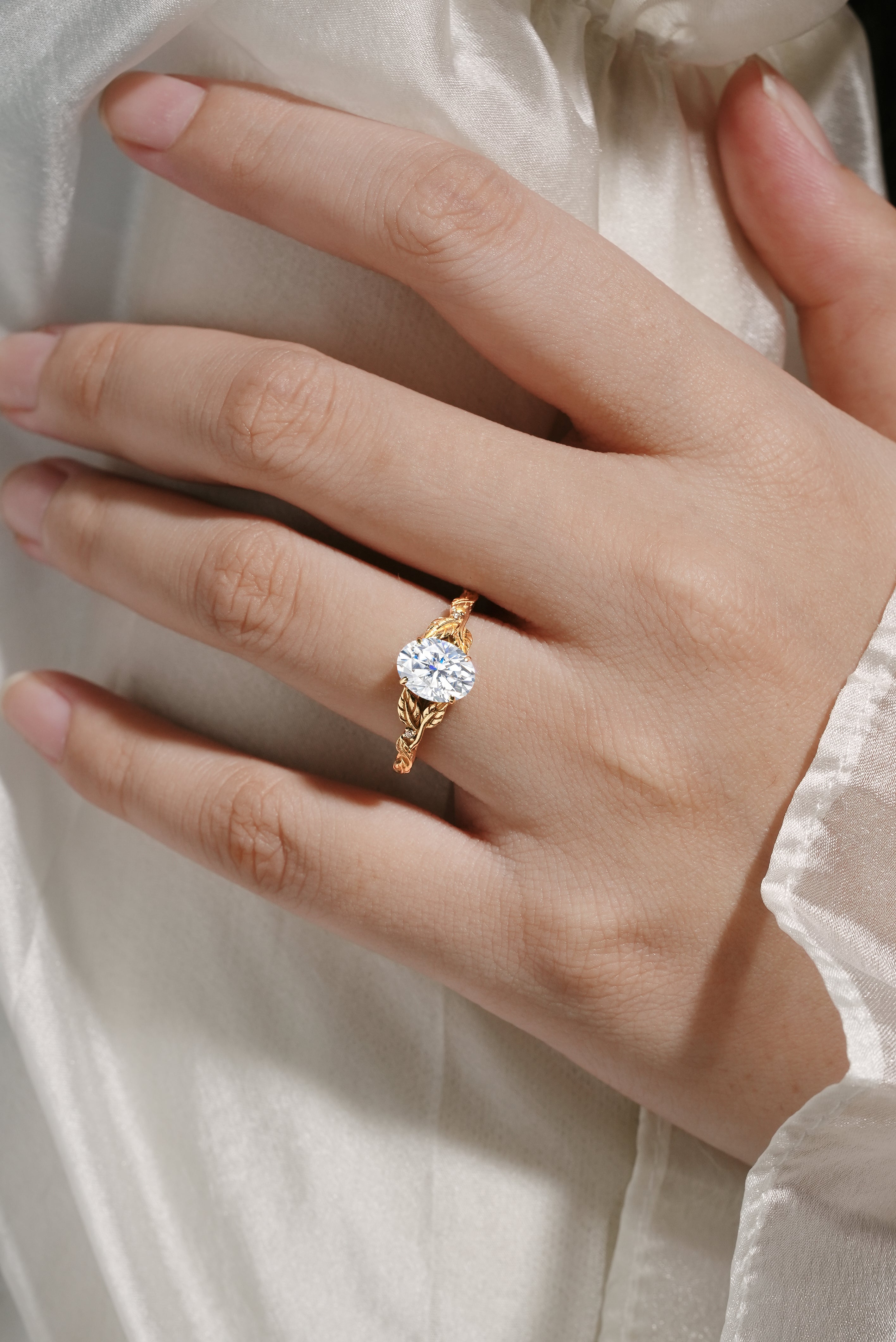 Oval moissanite engagement ring with leaf-inspired band on hand. 14k gold, bridal ring.